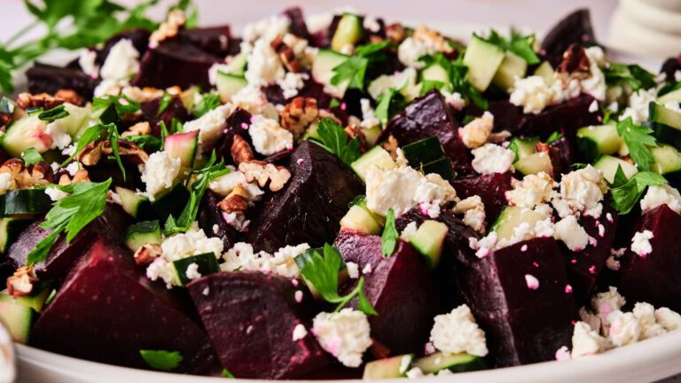 A close-up of a salad featuring chopped beets, cucumber, feta cheese, walnuts, and fresh herbs on a white plate.