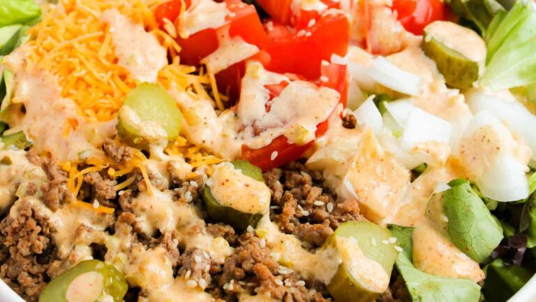A close-up of a salad topped with ground beef, shredded cheese, sliced pickles, chopped tomatoes, onions, and a creamy dressing.