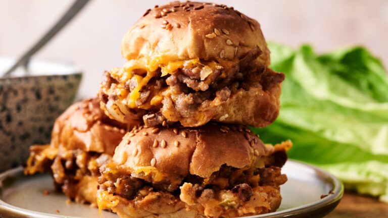 Two sesame seed buns filled with ground meat and melted cheese are stacked on a plate, with lettuce and a bowl in the background.