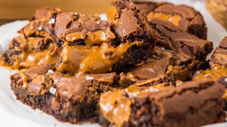 A pile of chocolate brownies with gooey caramel filling, topped with a sprinkle of coarse salt on a white surface.