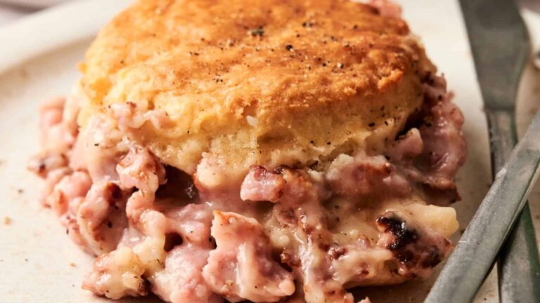 A biscuit topped with black pepper sits on a plate, filled with creamy diced ham gravy. A butter knife is placed beside it.
