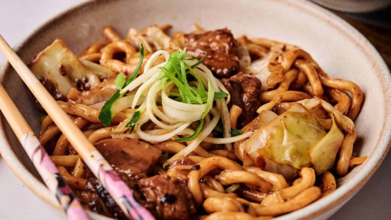 A bowl of stir-fried udon noodles with beef, cabbage, and green onions, served with chopsticks.