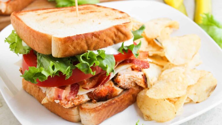 A sandwich with grilled chicken, bacon, lettuce, tomato, and a slice of cheese on toasted bread, served with potato chips on a white plate.