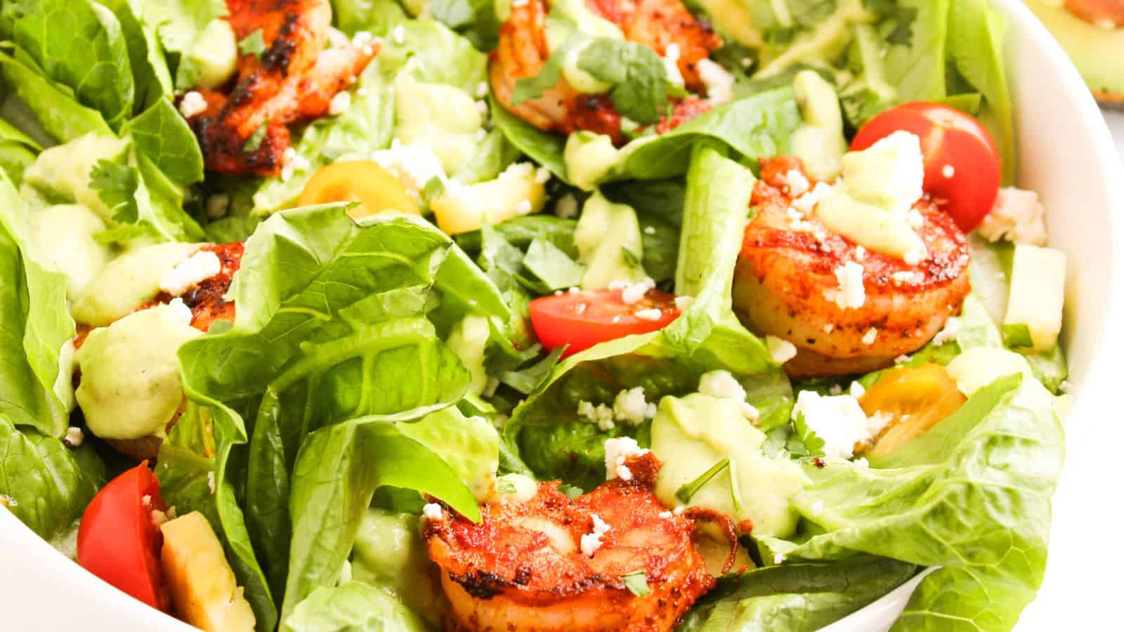A bowl of mixed salad with grilled shrimp, avocado, cherry tomatoes, lettuce, feta cheese, and a creamy dressing.