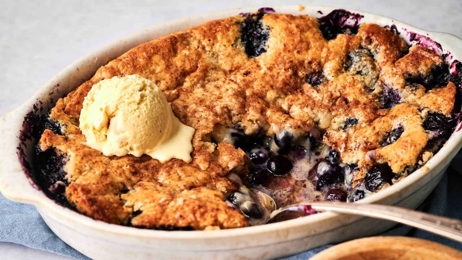 A baked blueberry cobbler with a golden crust is shown in a white dish, partially served, topped with a scoop of vanilla ice cream.