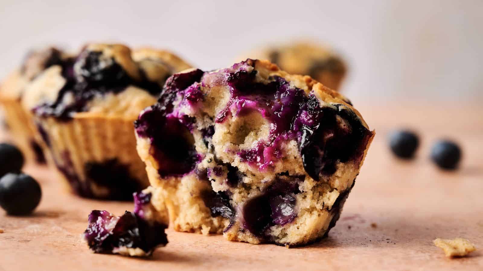 A close-up of a blueberry muffin with a bite taken out, showing the moist interior and whole blueberries; other muffins and loose blueberries are in the background.