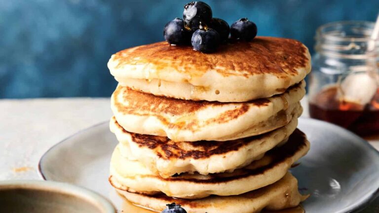 A stack of pancakes topped with blueberries and syrup sits on a plate, with a jar of syrup in the background.