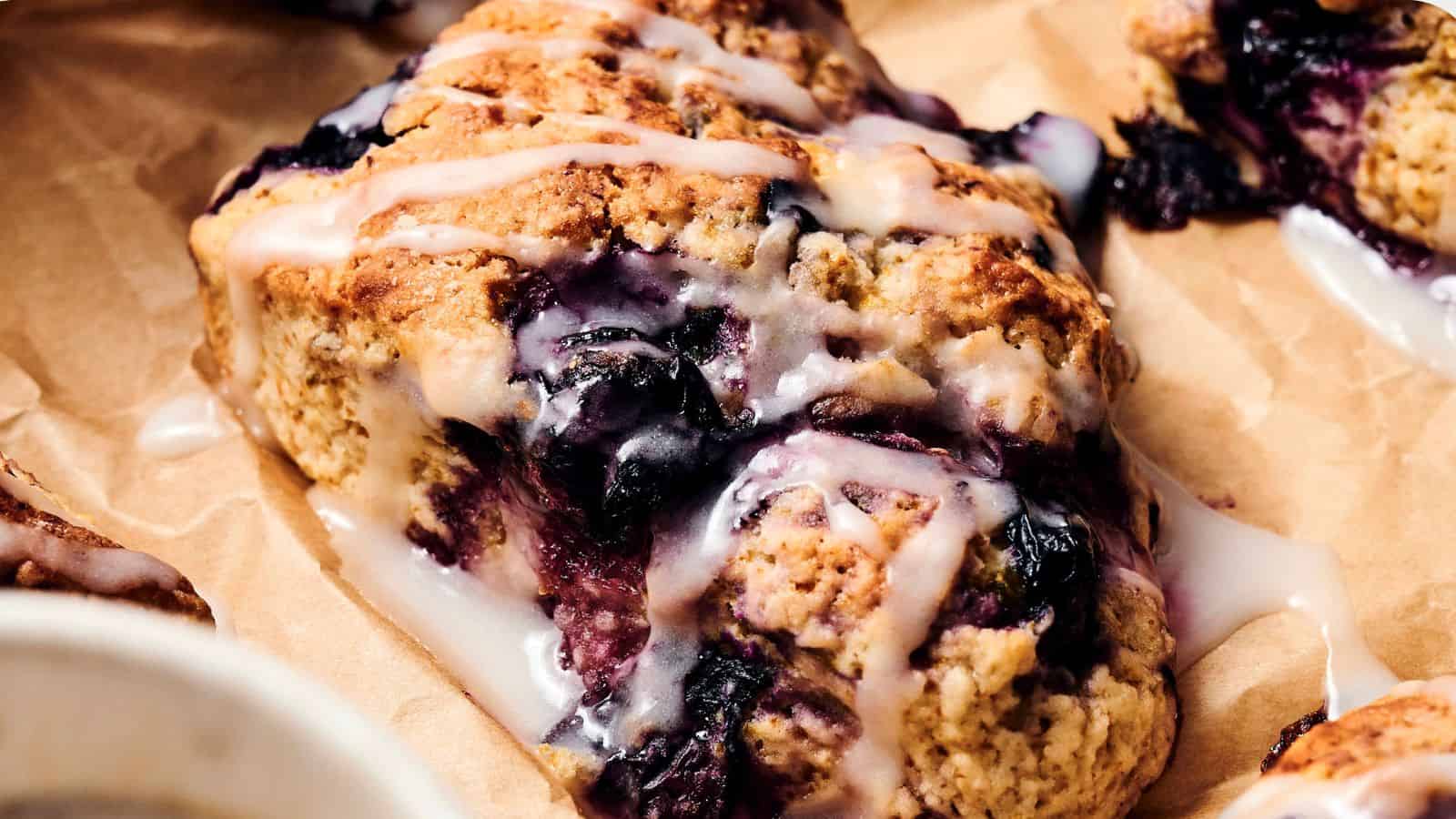 A close-up of a blueberry scone with a light glaze, resting on brown parchment paper.