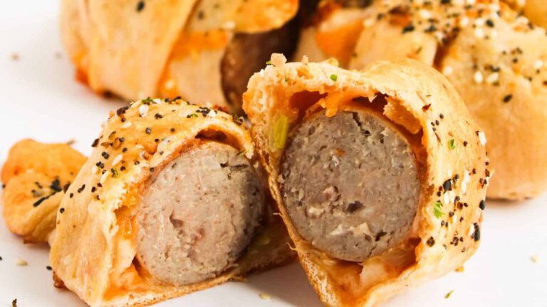 A close-up of sliced sausage wrapped in golden-brown pastry, topped with seasoning and visible herbs.