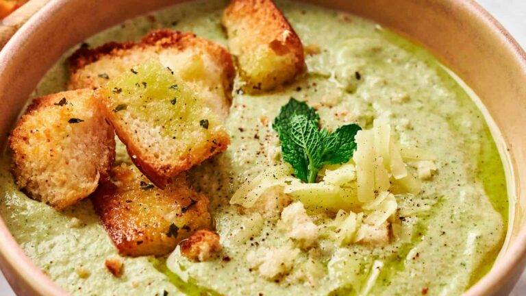 A close-up of creamy green soup garnished with croutons, shredded cheese, fresh mint, cracked pepper, and a drizzle of olive oil in a bowl.