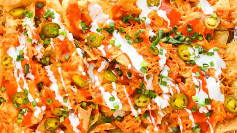 Close-up of nachos topped with shredded chicken, sliced jalapeños, hot sauce, and drizzles of sour cream, garnished with chopped green onions.
