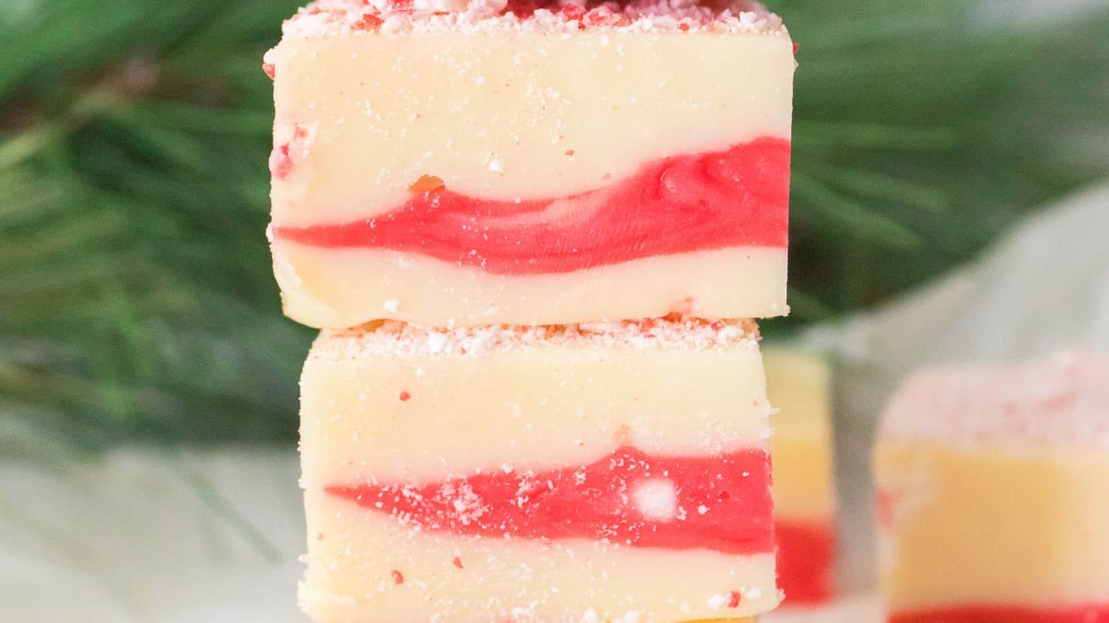 Two pieces of vanilla and red swirled candy cane fudge stacked, with a sprinkle of crushed peppermint on top. Greenery in the background.