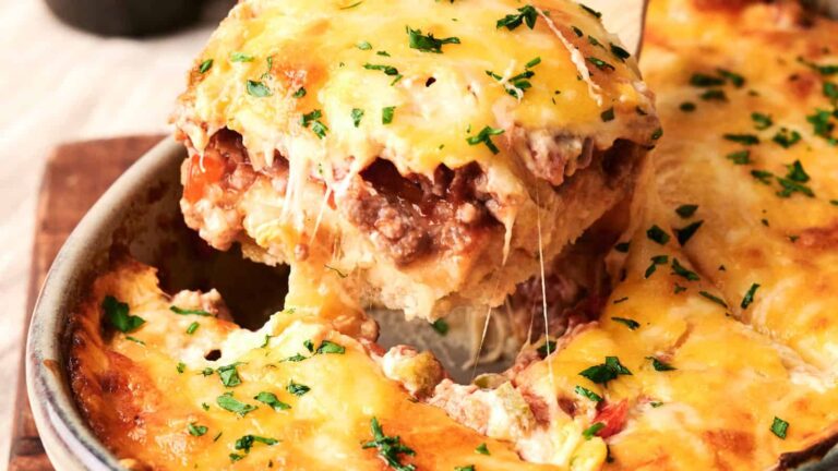 A close-up of a cheesy baked lasagna being lifted from a pan, topped with melted cheese and chopped herbs.
