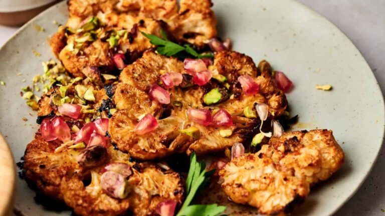 Roasted cauliflower steaks garnished with pomegranate seeds, chopped pistachios, and fresh parsley on a light-colored plate.
