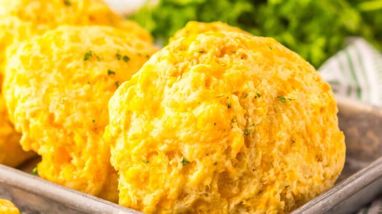 Close-up of golden, cheesy biscuits on a baking tray with a garnish of herbs, with leafy greens blurred in the background.
