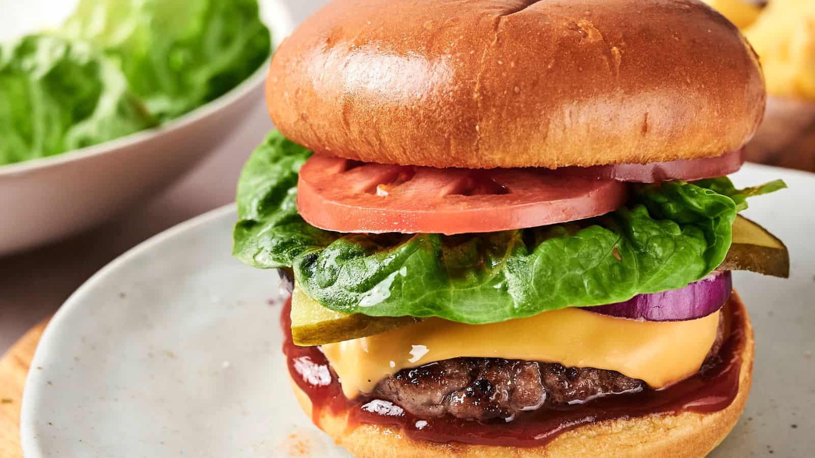 A cheeseburger with lettuce, tomato, pickles, onion, and ketchup on a plate. A bowl of lettuce is in the background.