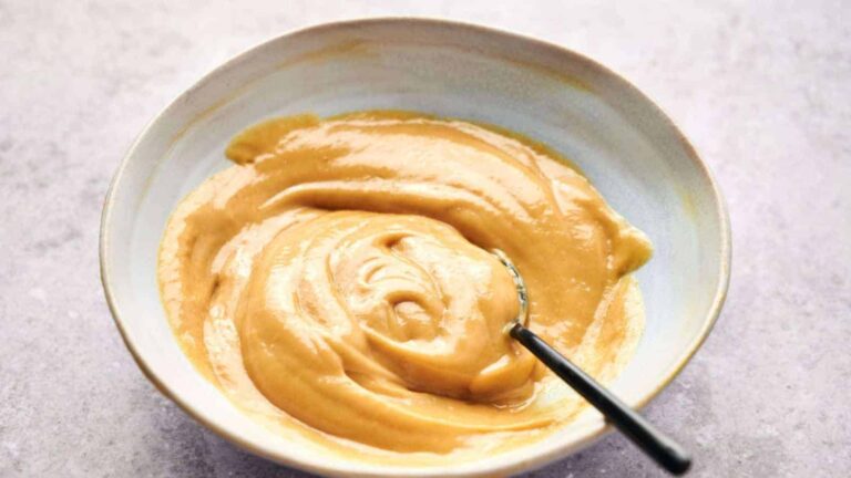 A bowl filled with creamy, light brown sauce or paste, with a spoon partially submerged in the mixture.