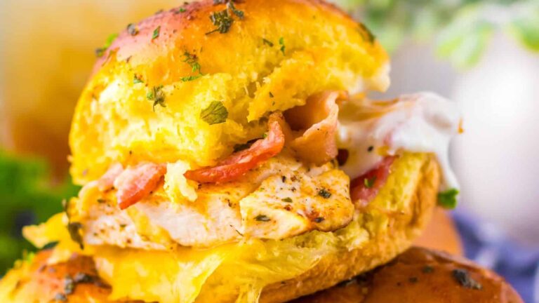 A close-up of a sandwich with grilled chicken, crispy bacon, melted cheese, and herbs on a golden bun.