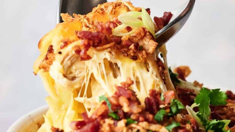 A close-up of a cheesy pasta dish topped with crispy bacon, green onions, and herbs, with a spoon lifting a serving and melted cheese stretching.