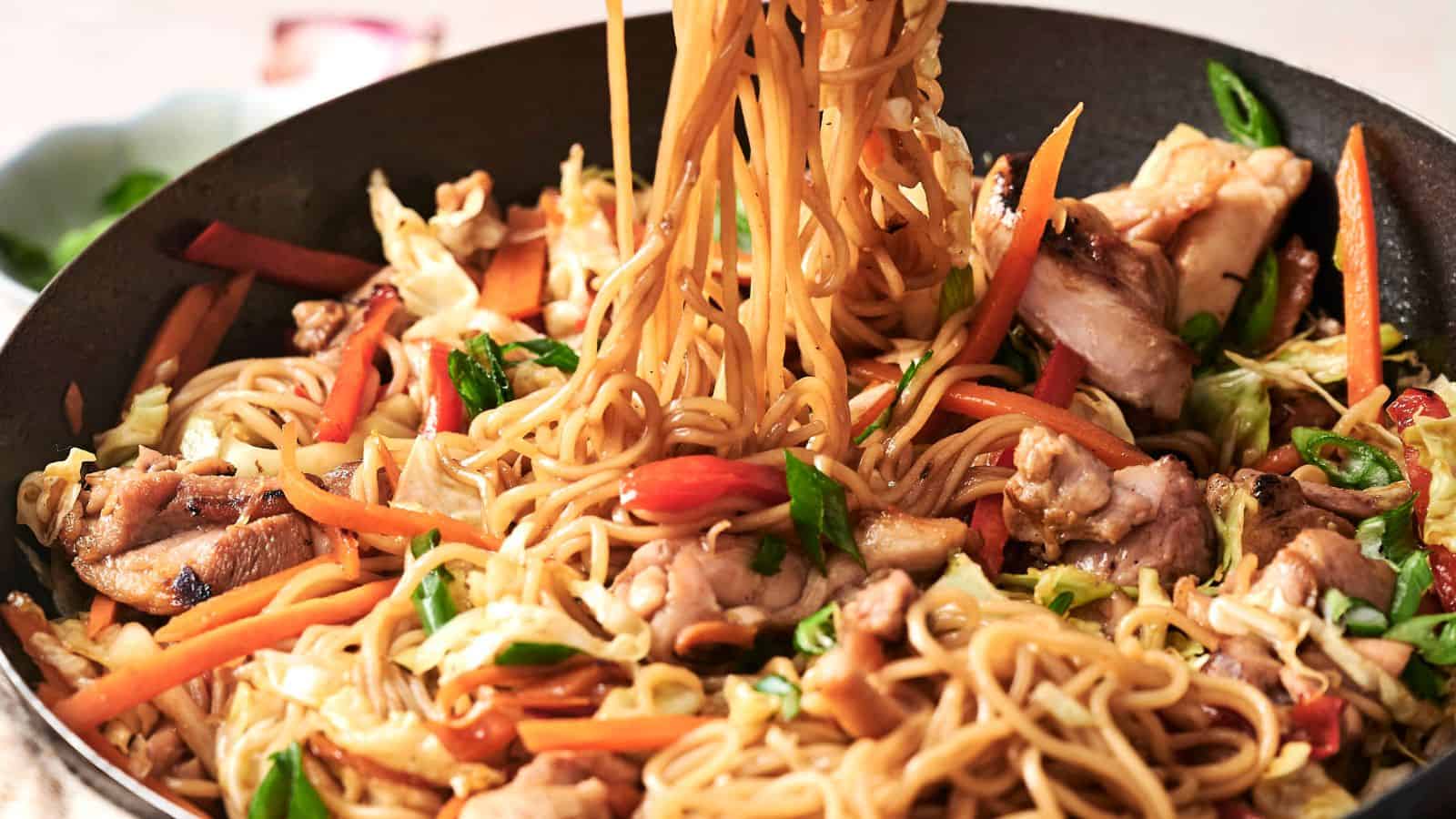 Stir-fried noodles with vegetables and chicken in a pan, featuring carrots, cabbage, and green onions.