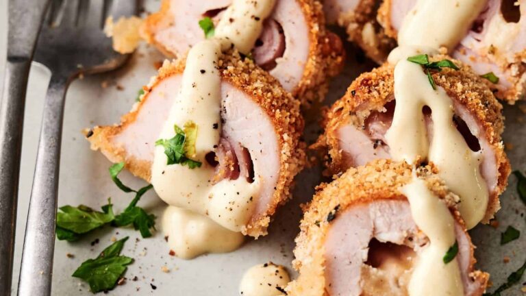 Sliced breaded chicken stuffed with ham and cheese, topped with creamy sauce and garnished with chopped herbs on a plate.