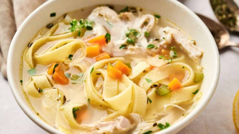 A bowl of chicken noodle soup with wide egg noodles, chunks of chicken, carrots, celery, and parsley in a clear broth.