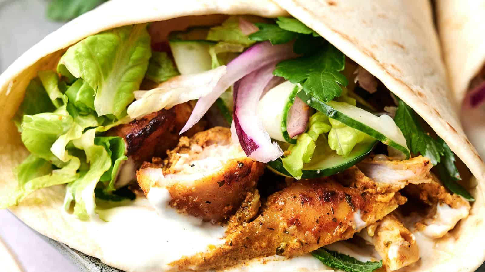 Close-up of a wrap filled with grilled chicken, lettuce, cucumber, red onion, and sauce in a folded flatbread.