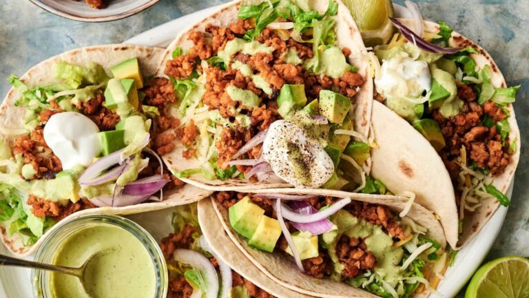 Three soft tacos filled with seasoned ground meat, shredded lettuce, diced avocado, red onion, shredded cheese, and dollops of sour cream, served with a green sauce on the side.