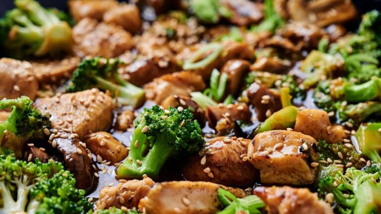 Close-up of stir-fried chicken, broccoli, and mushrooms in a savory sauce, topped with sesame seeds.