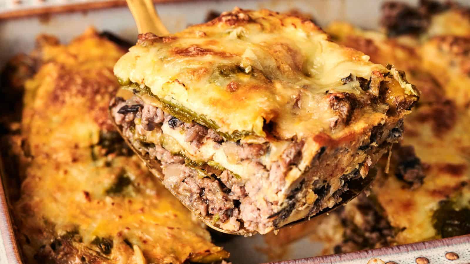 A slice of lasagna with beef, mushrooms, cheese, and vegetables being lifted from a baking dish.