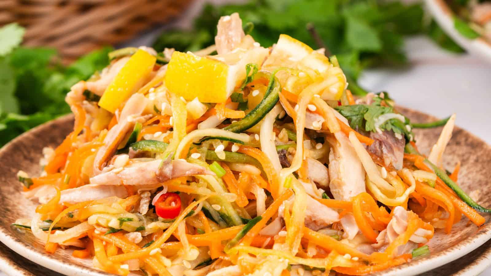 A colorful salad with shredded carrots, zucchini, chicken, orange slices, and sesame seeds on a brown plate, garnished with fresh herbs.