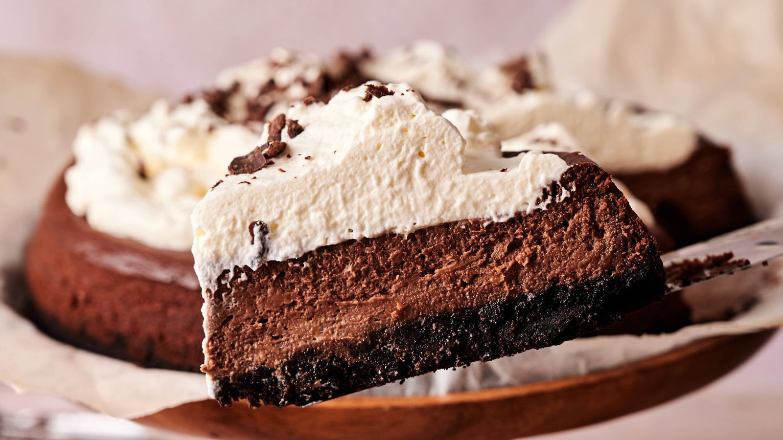 A slice of chocolate cheesecake with a cookie crust and whipped cream topping is being lifted from the whole cake.