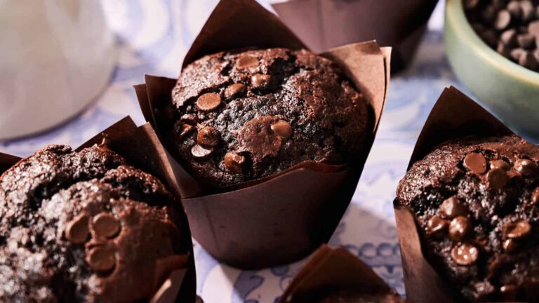 Close-up of chocolate chip muffins in brown paper wrappers, with more chocolate chips visible in a bowl in the background.