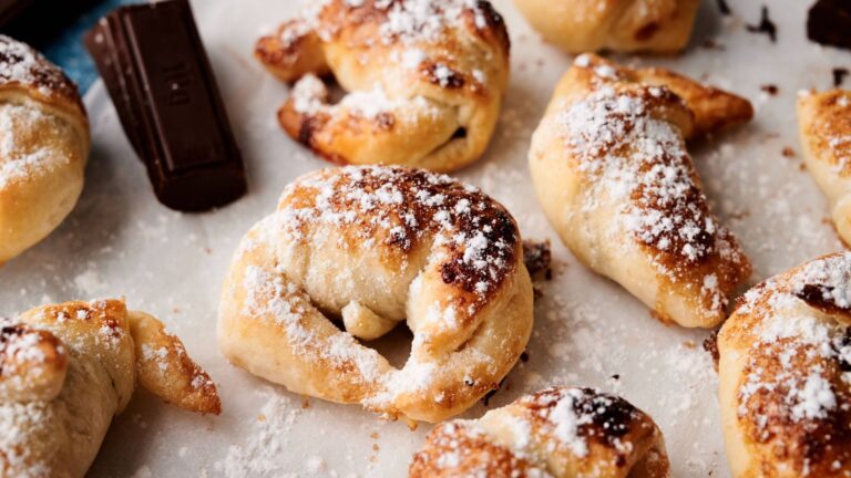 Close-up of baked pastries dusted with powdered sugar on parchment paper, with pieces of chocolate nearby.