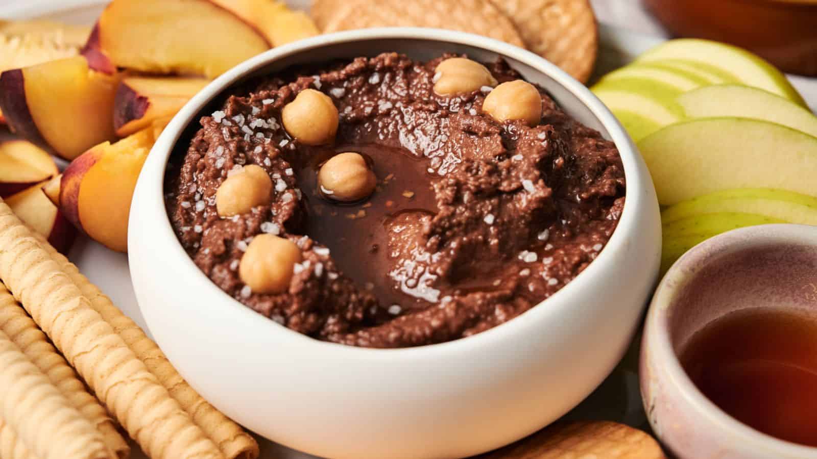 A bowl of chocolate hummus topped with chickpeas and coarse salt, surrounded by sliced peaches, apples, biscuits, and rolled wafers.