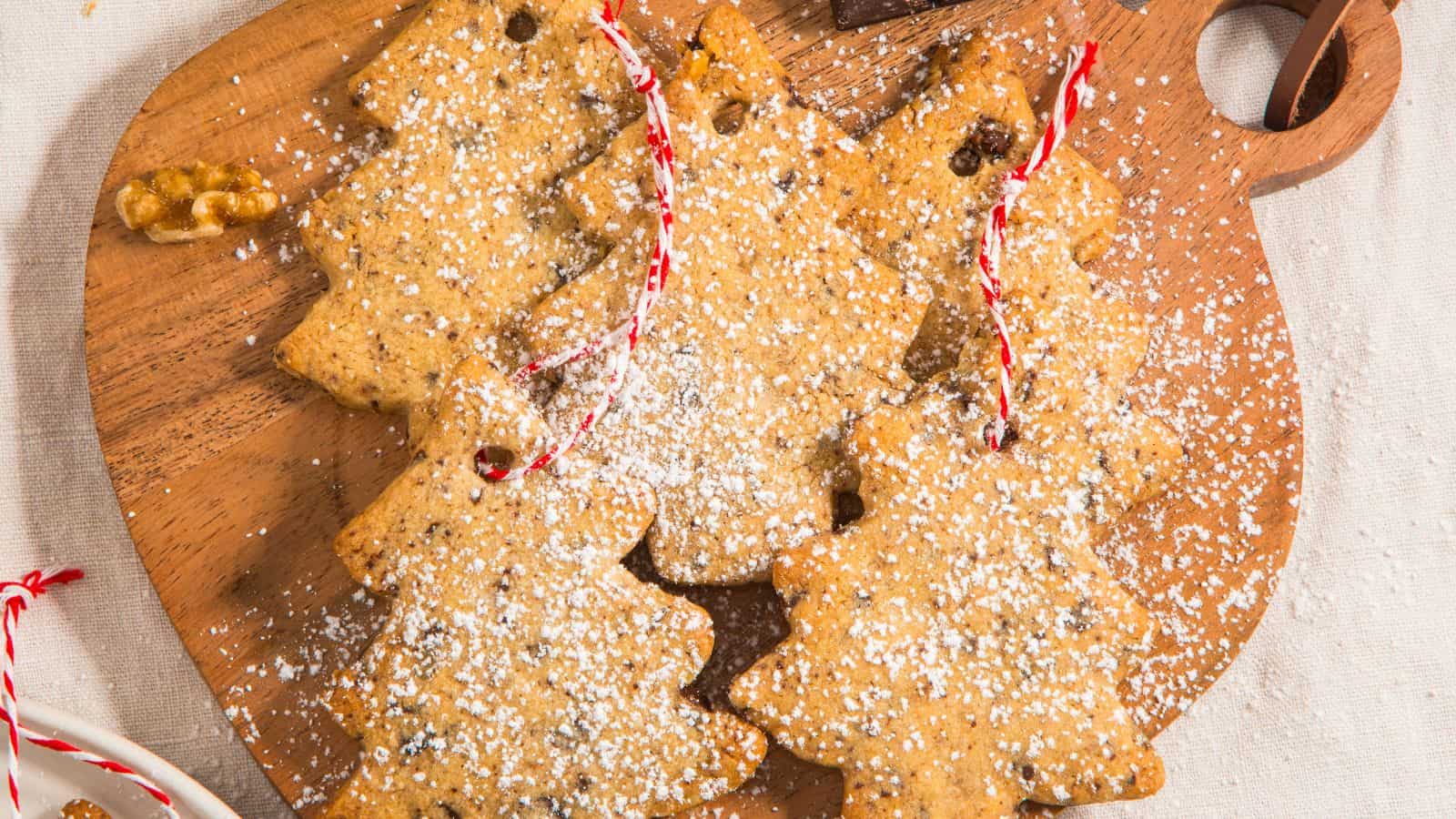 Four Christmas tree-shaped cookies with powdered sugar on a wooden board, decorated with red and white string.