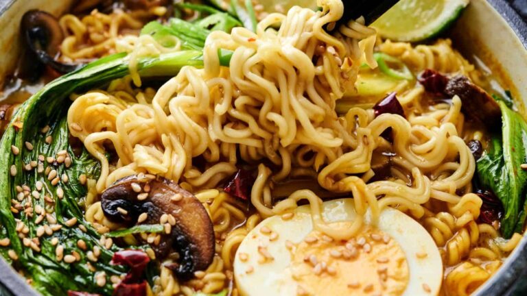 A bowl of ramen noodles with mushrooms, bok choy, a halved boiled egg, lime slices, sesame seeds, and chopsticks lifting noodles.
