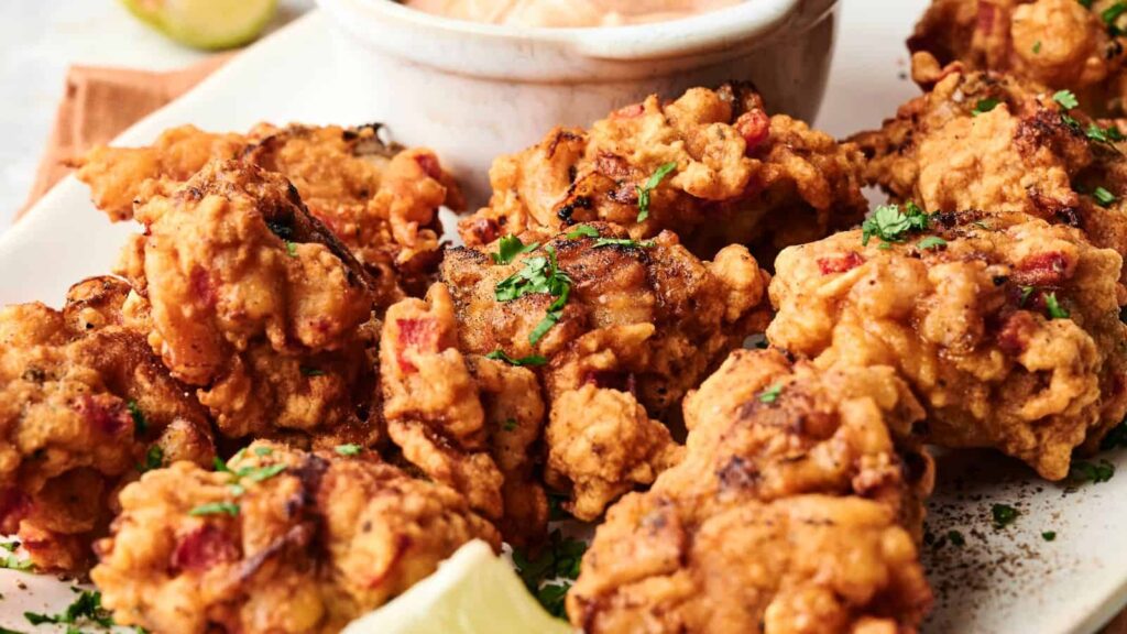 A plate of golden-brown fritters garnished with chopped herbs, served with a dipping sauce and a wedge of lemon.