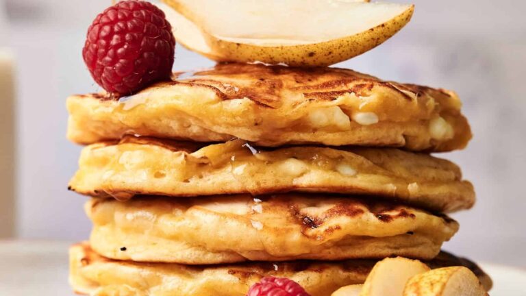 A stack of three pancakes topped with a pear slice and raspberries, with syrup drizzled over them.