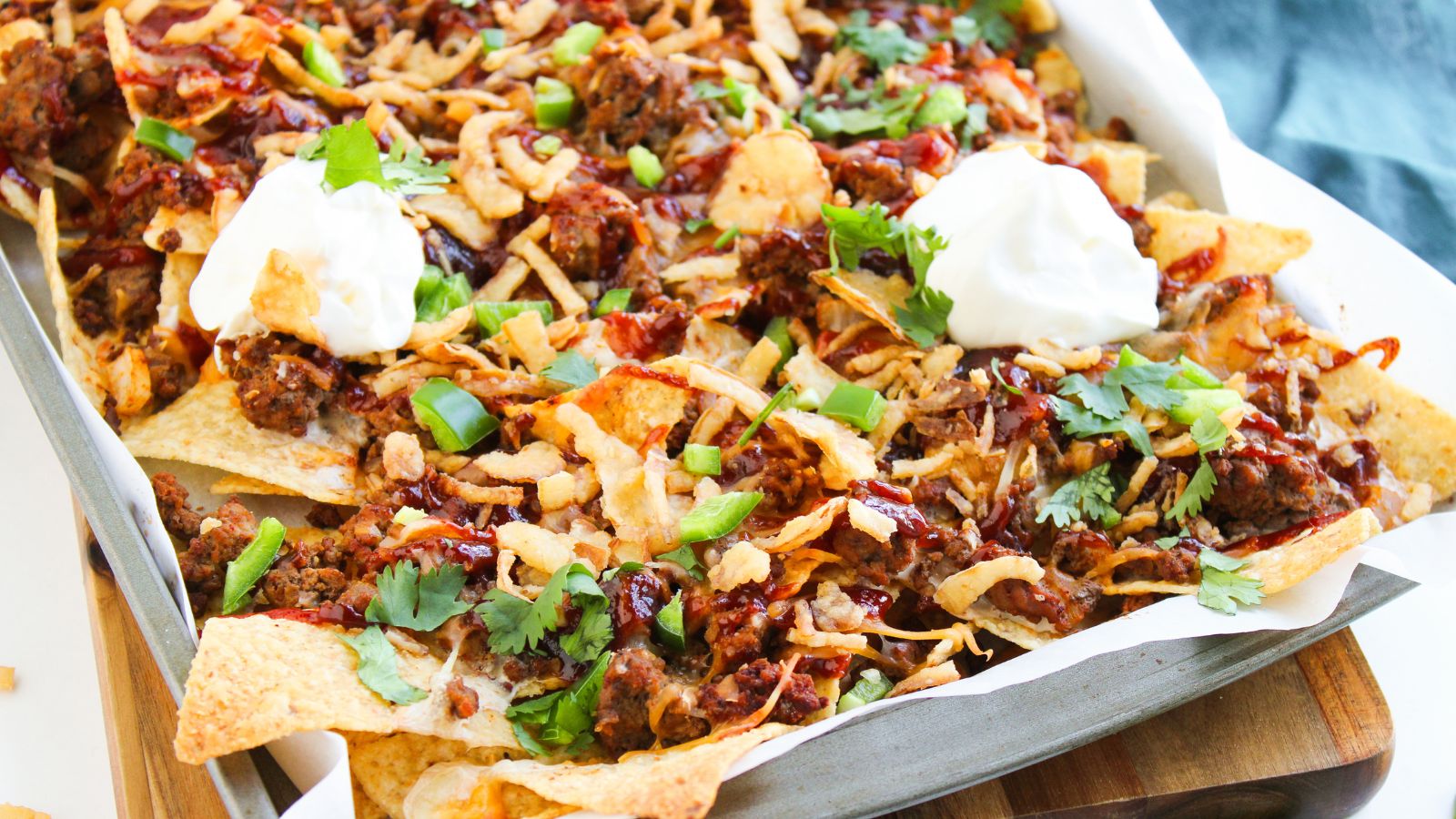 A tray of nachos topped with ground meat, melted cheese, chopped green onions, cilantro, fried onions, barbecue sauce, and two dollops of sour cream.