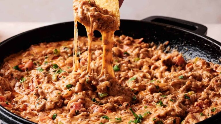 A skillet filled with cheesy, ground meat dip topped with herbs, with a tortilla chip scooping up a portion of the dip.