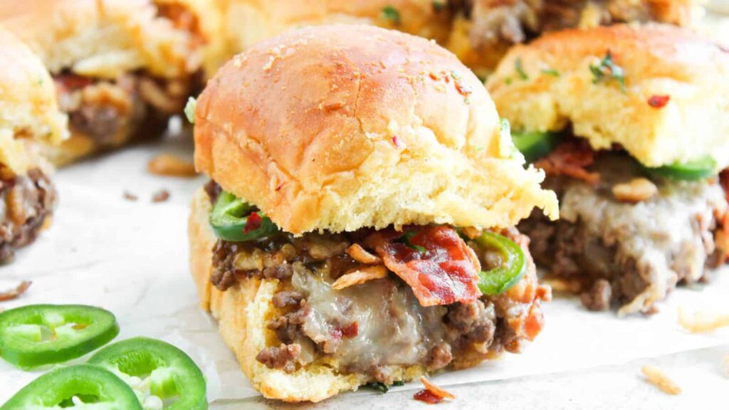 A close-up of sliders filled with cooked ground beef, melted cheese, bacon, and sliced jalape&ntilde;os on a white surface, with some jalape&ntilde;o slices scattered nearby.