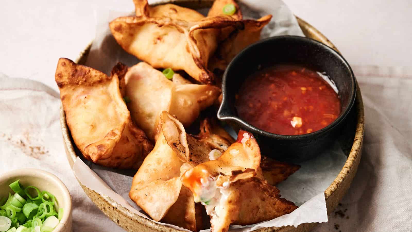 A bowl of fried wontons served with a side of red dipping sauce and a small dish of chopped green onions.