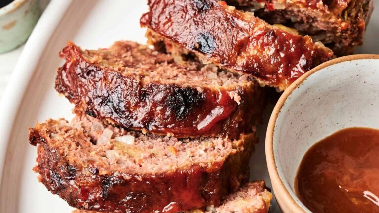 Three slices of glazed meatloaf are arranged on a white plate next to a small bowl of brown sauce.