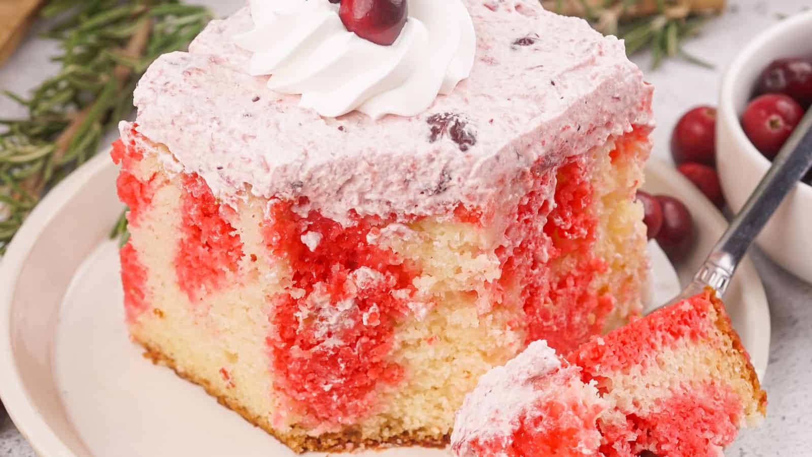 A slice of festive Christmas Poke Cake with red swirls and pink frosting, topped with a dollop of whipped cream and a cherry, sits elegantly on a plate with a fork.