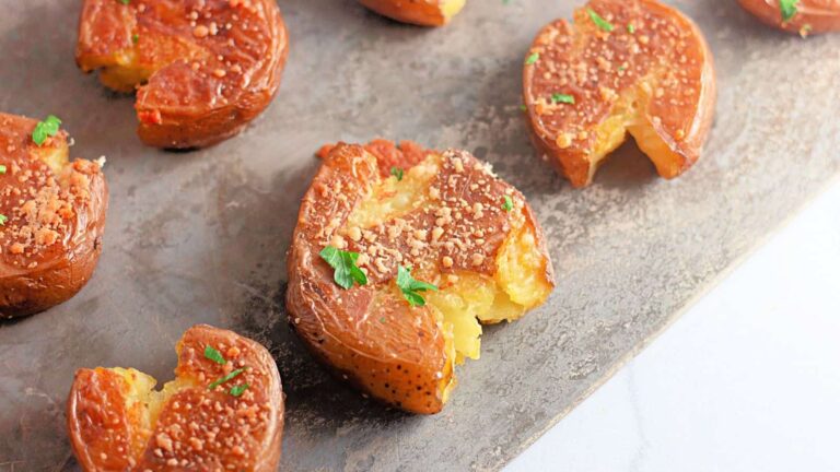 Several smashed roasted potatoes topped with grated cheese and chopped parsley are arranged on a metal surface.