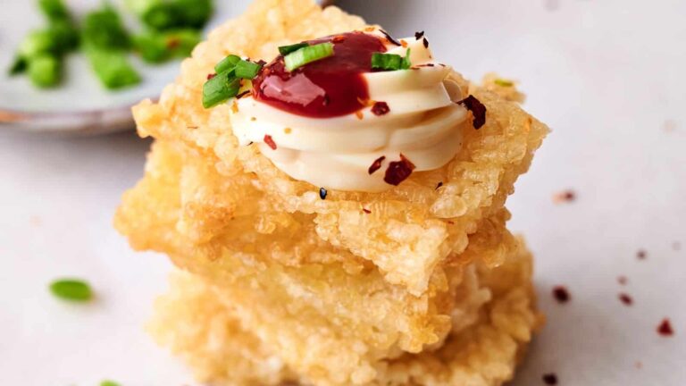 Two pieces of crispy fried tofu stacked, topped with a dollop of mayonnaise, red sauce, chopped green onions, and red pepper flakes.