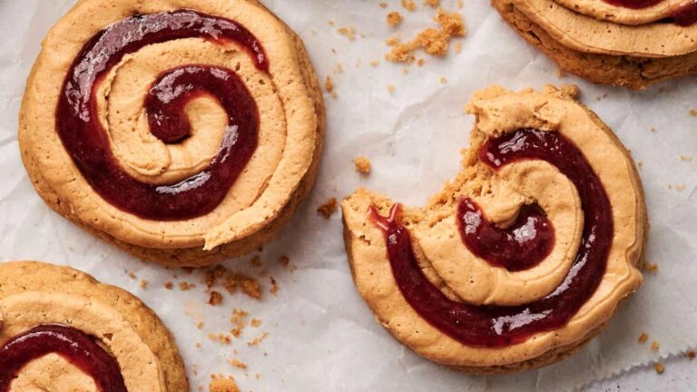 Three peanut butter and jelly swirl cookies on parchment paper, with one cookie partially eaten and crumbs scattered around.