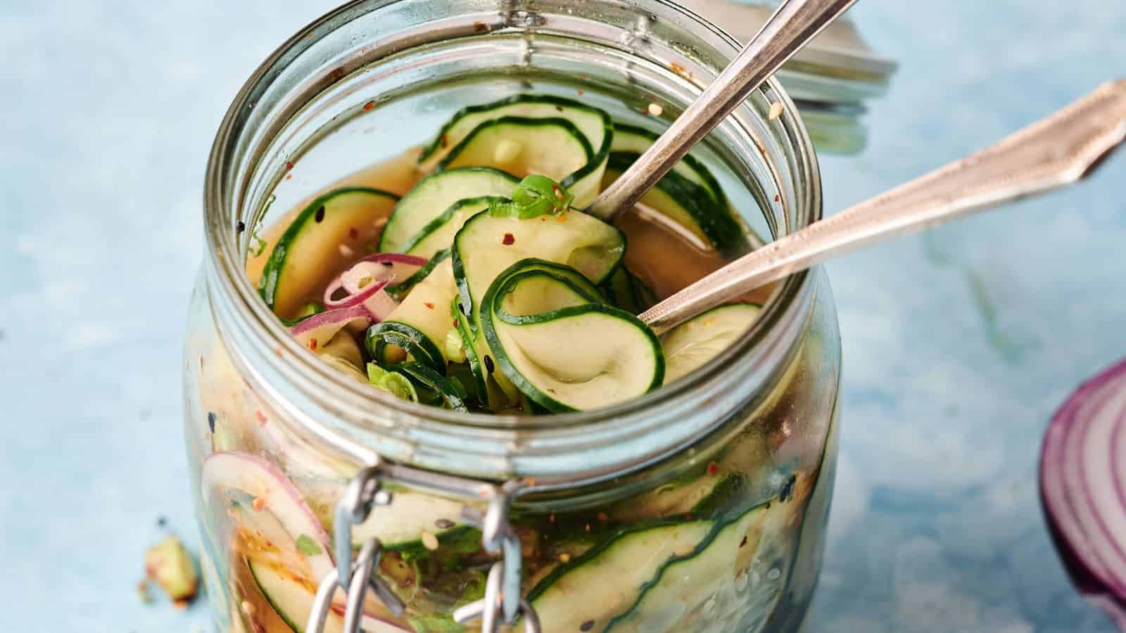 A glass jar filled with sliced cucumbers, red onions, and herbs in pickling liquid, with two metal forks placed inside.
