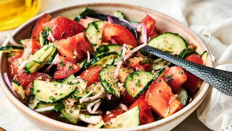 A bowl of fresh salad with chopped tomatoes, cucumbers, red onions, and herbs, mixed and topped with black pepper, with a spoon resting inside.